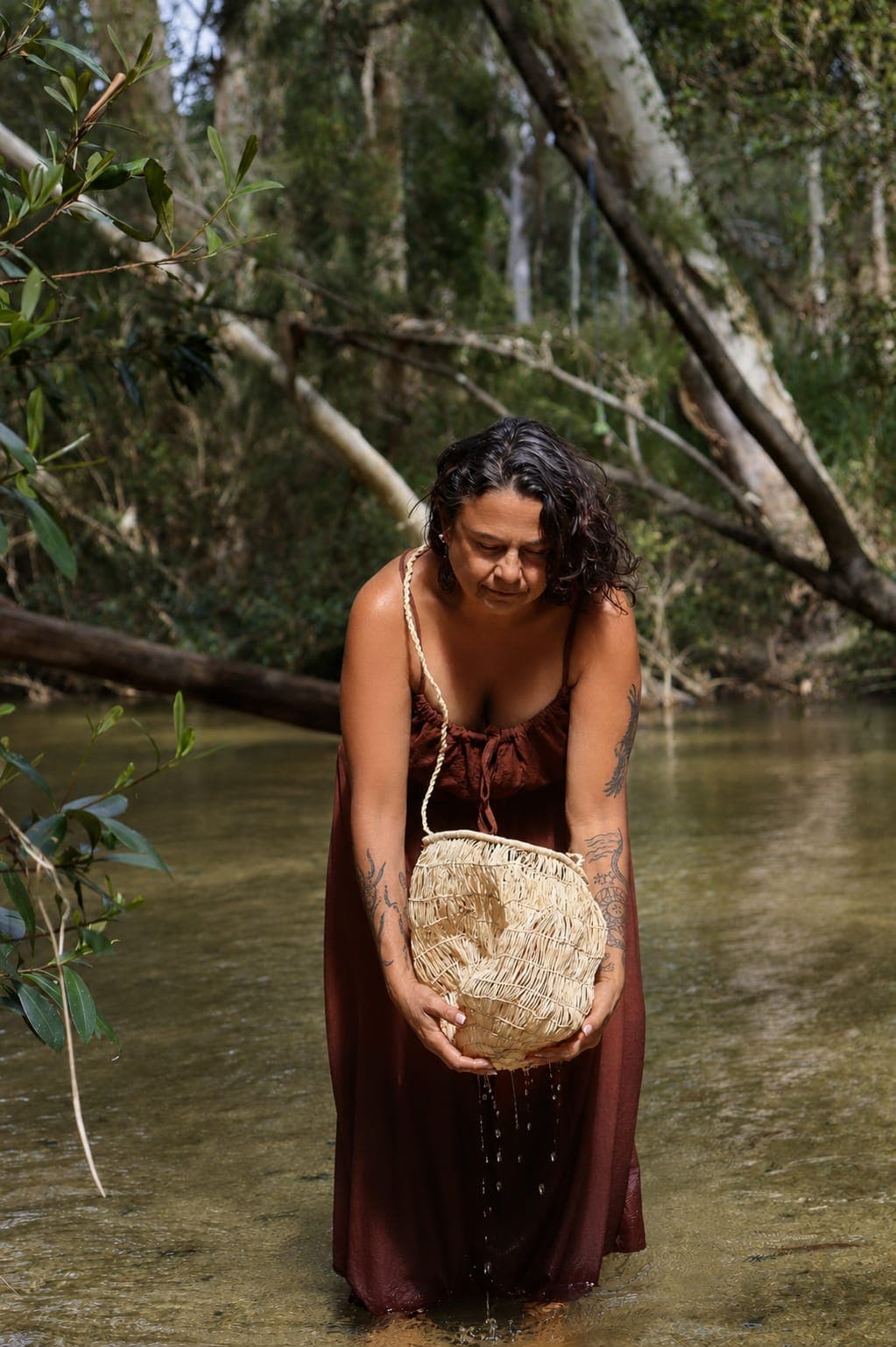 Julie standing in a shallow creek on Country with a woven fibre bag.